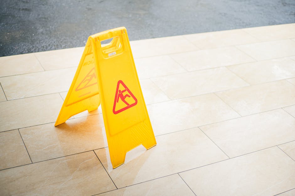A yellow plastic warning sign with a red triangle and a black icon of a person slipping is placed on a smooth, beige tiled floor outdoors, indicating a wet or slippery surface. The sign stands upright